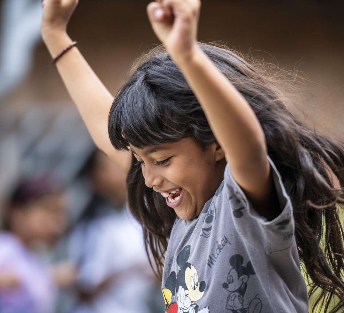 Nahaufnahme eines fröhlichen Mädchens mit langen dunklen Haaren, die um sie herumfliegen, lachend mit geschlossenen Augen und erhobenen Armen in einem grauen Micky-Maus-Shirt.