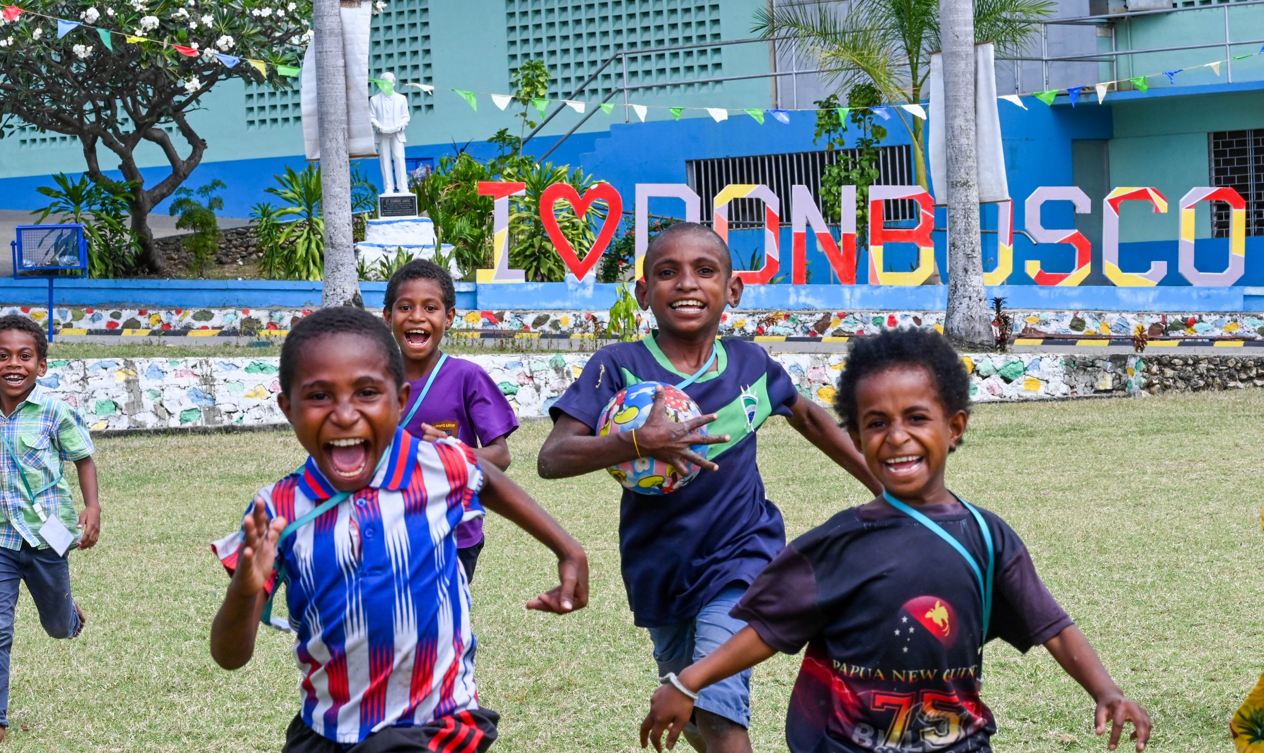 Fröhliche Kinder rennen auf einem Rasenfeld vor einem farbigen "I LOVE DON BOSCO" Schild.