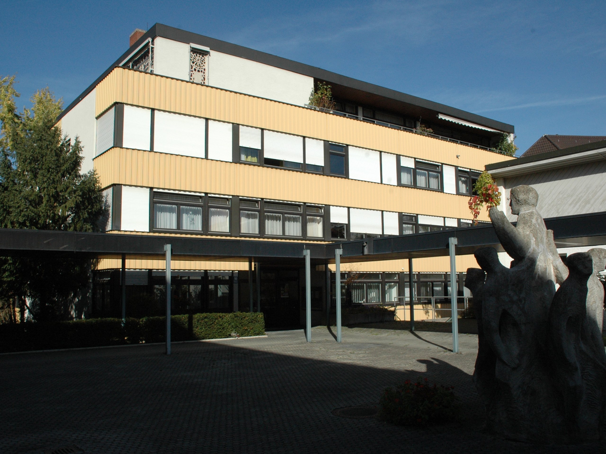 Ein modernes gelb-weißes Gebäude, ein Carport, Steinskulpturen und ein großer Baum unter klarem blauem Himmel.