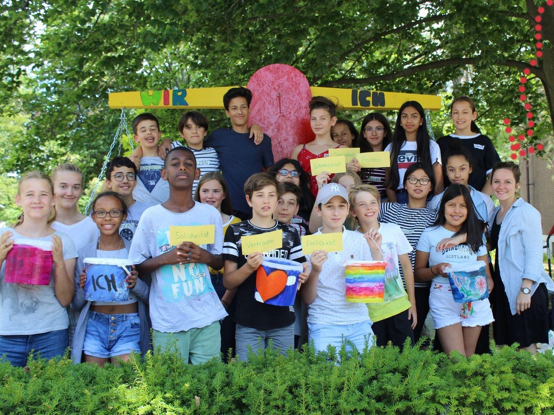 Vielfältige Jugendliche und eine Erwachsene posieren draußen vor einem 'WIR'- und 'ICH'-Schild und halten Karten mit Werten.