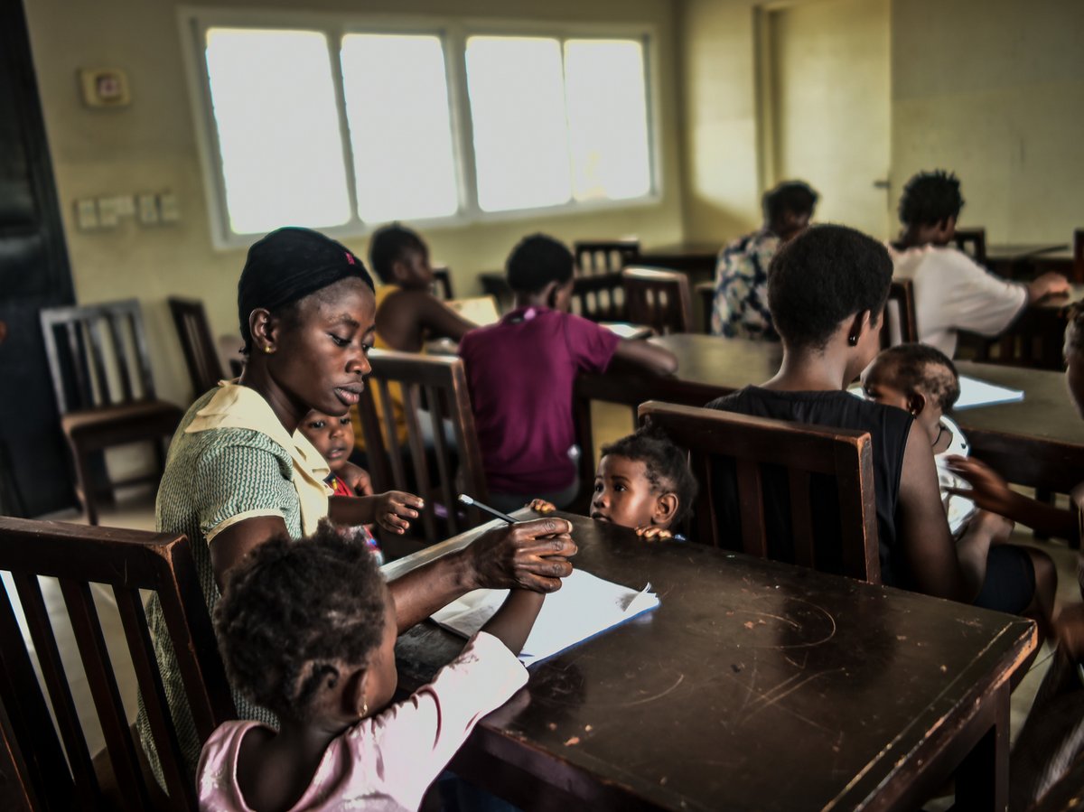 Eine Frau hilft Kindern an einem Holztisch beim Lernen, umgeben von anderen Erwachsenen und Kindern. Ein Kind sitzt auf ihrem Schoß.