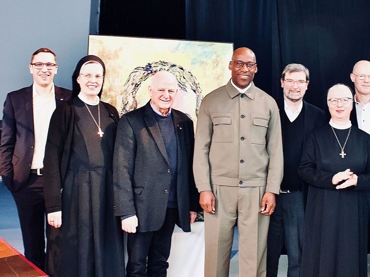 Sieben lächelnde Personen, darunter zwei Nonnen und fünf Männer, posieren gemeinsam vor einem abstrakten Gemälde bei einer Veranstaltung.