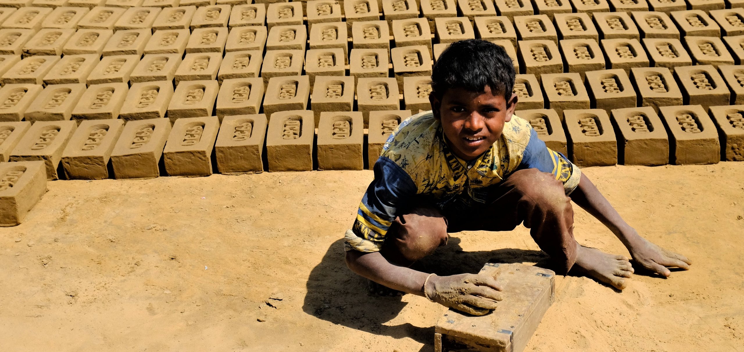 Ein Junge, schmutzig von der Ziegelherstellung, hockt auf dem Boden, umgeben von Reihen trocknender Lehmziegel.
