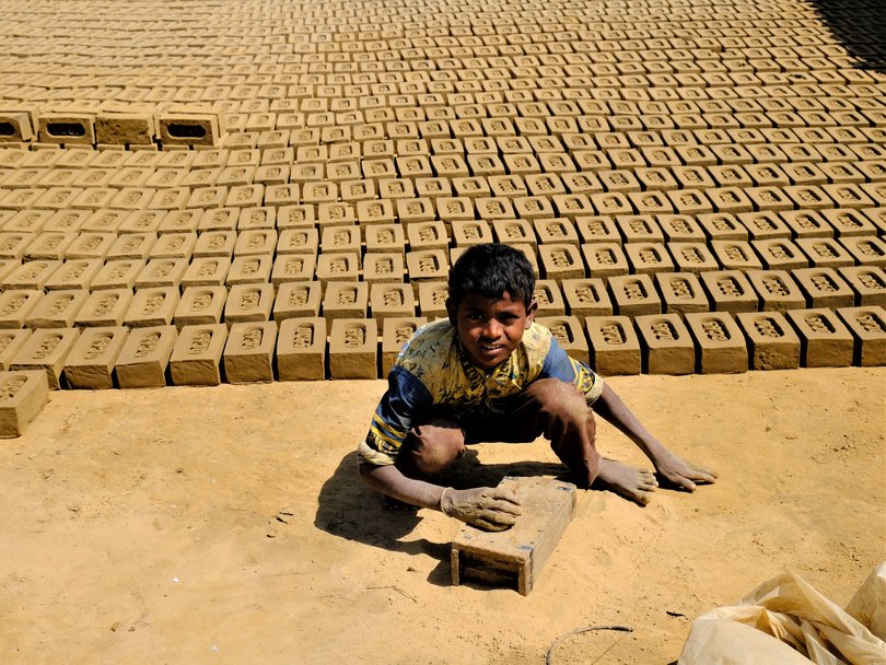 Ein Junge, schmutzig von der Ziegelherstellung, hockt auf dem Boden, umgeben von Reihen trocknender Lehmziegel.
