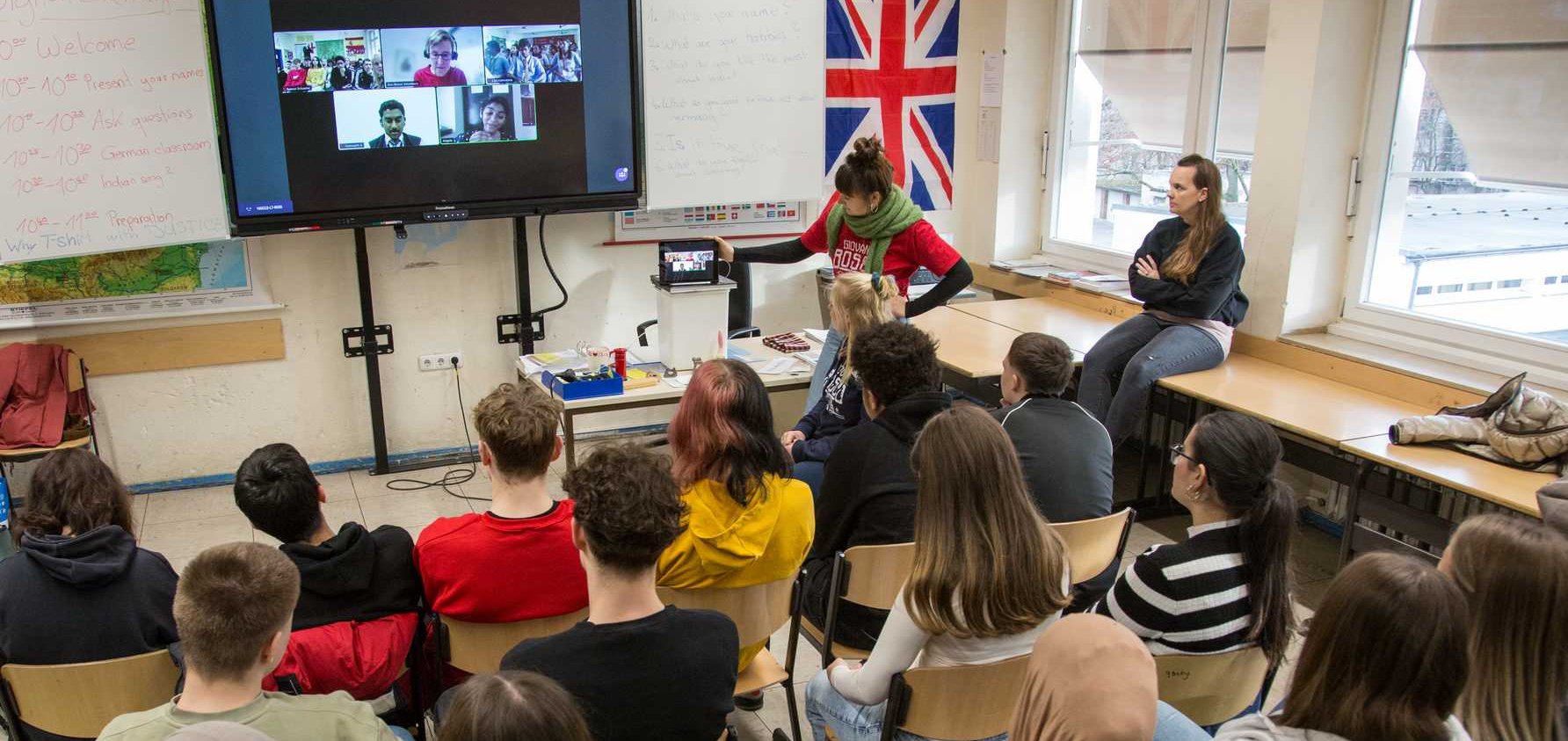 Schüler in einem Klassenzimmer nehmen an einem digitalen Austausch per Videokonferenz teil, eine Lehrerin moderiert.