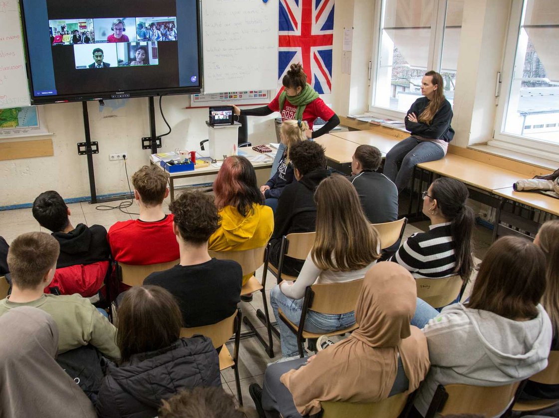 Schüler in einem Klassenzimmer nehmen an einem digitalen Austausch per Videokonferenz teil, eine Lehrerin moderiert.