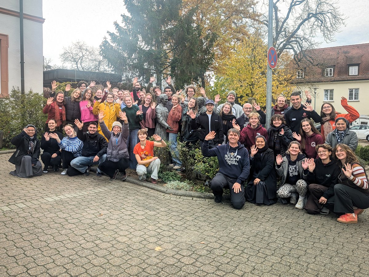 Große Gruppe lachender, winkender Menschen verschiedenen Alters im Freien vor Gebäuden und bunten Herbstbäumen.