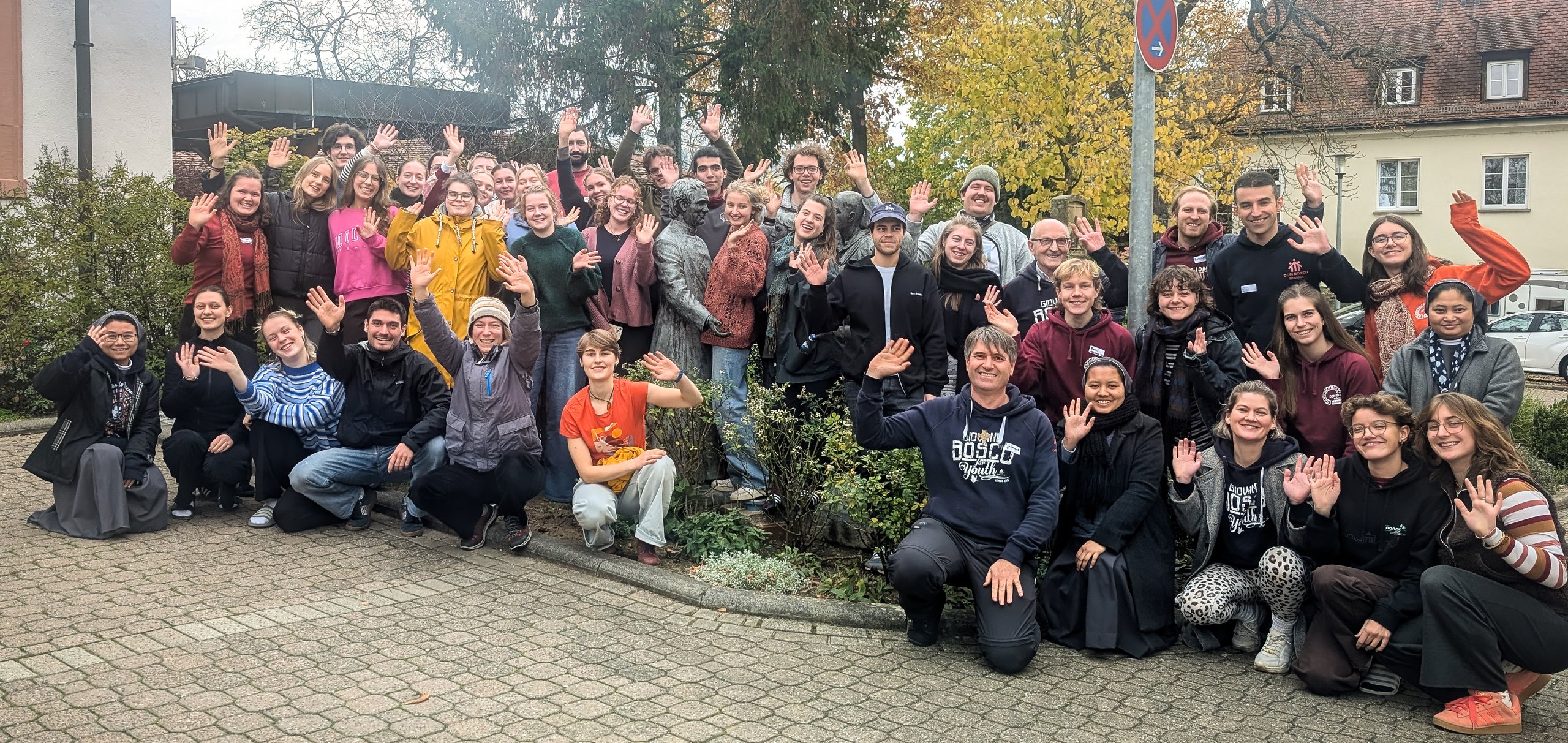 Eine große Gruppe lächelnder junger und älterer Erwachsener posiert im Freien, viele winken. Herbstliche Bäume und Gebäude im Hintergrund.