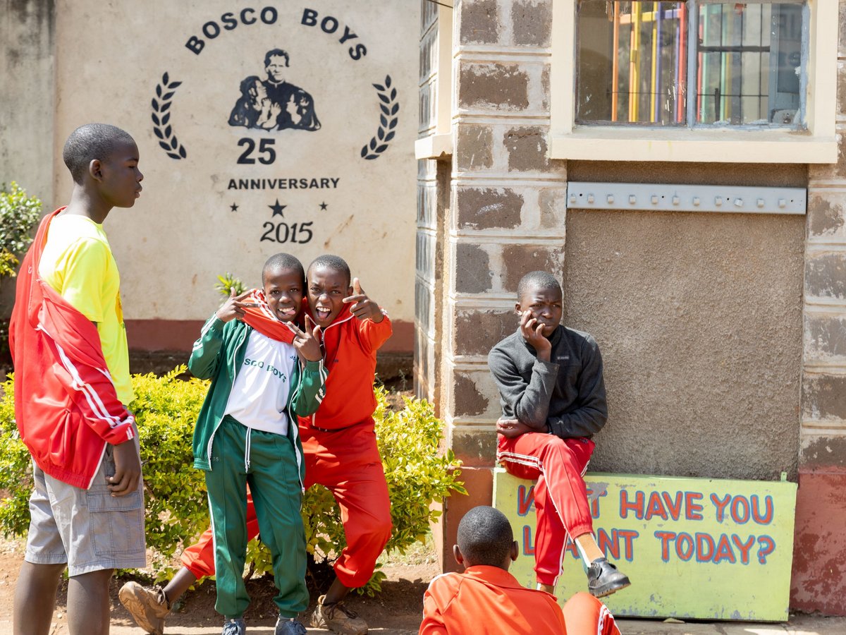 Vier Jungen vor einem 'Bosco Boys' Gebäude. Zwei posieren spielerisch für die Kamera, einer schaut zu, ein anderer sitzt nachdenklich.