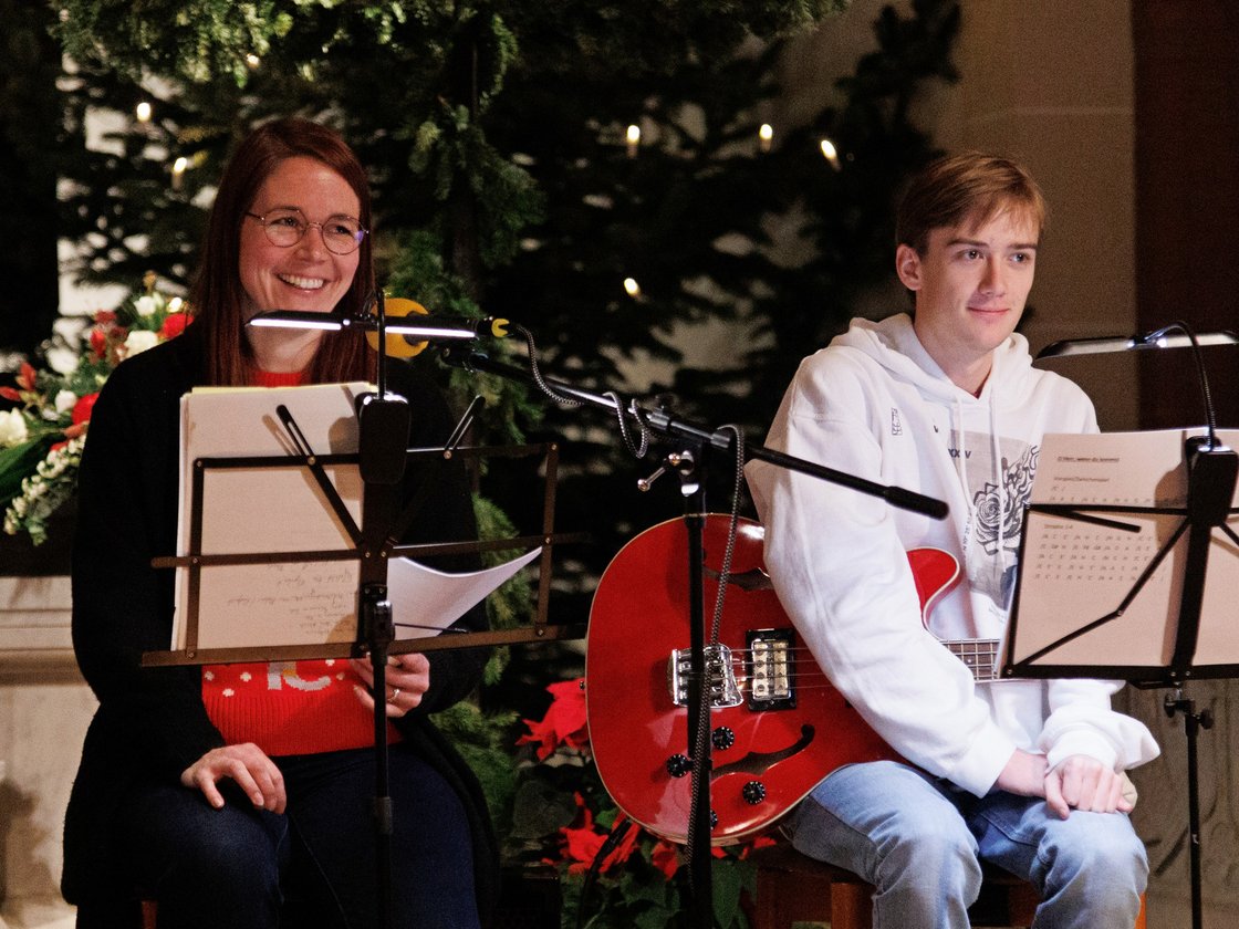 Zwei Musiker, eine lächelnde Frau und ein junger Mann mit roter Bassgitarre, sitzen vor festlichem Grün mit Notenständern.