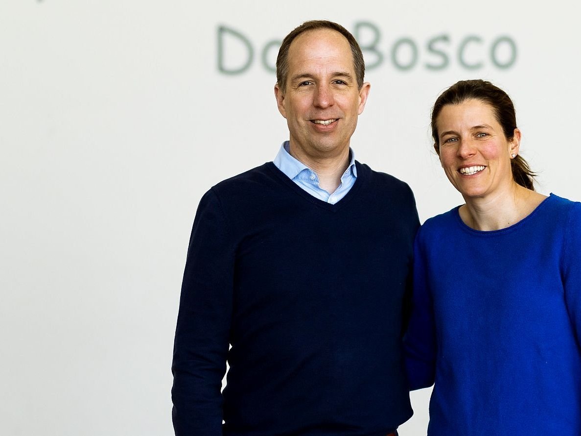 Ein lächelnder Mann im blauen Pullover und eine Frau im blauen Oberteil stehen nebeneinander vor einer weißen Wand mit 'Do Bosco'-Schriftzug.
