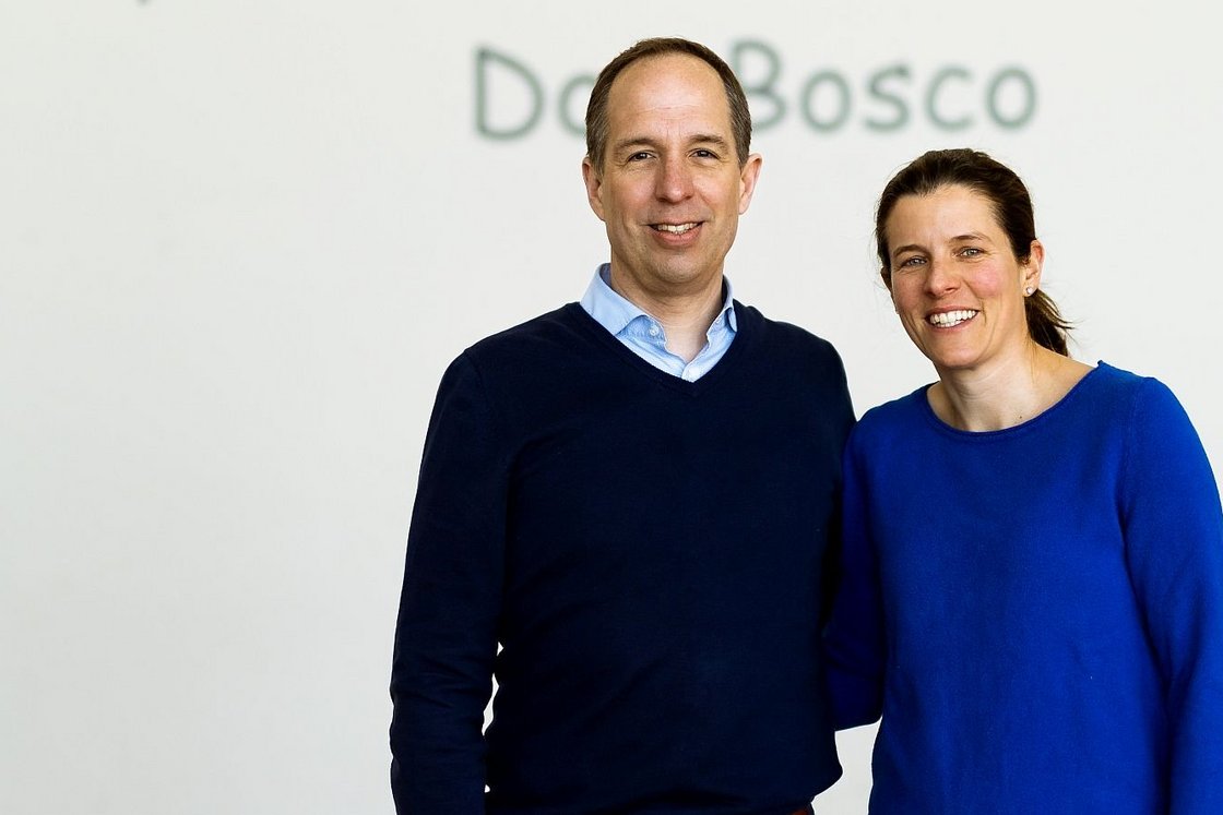 Ein lächelnder Mann im blauen Pullover und eine Frau im blauen Oberteil stehen nebeneinander vor einer weißen Wand mit 'Do Bosco'-Schriftzug.
