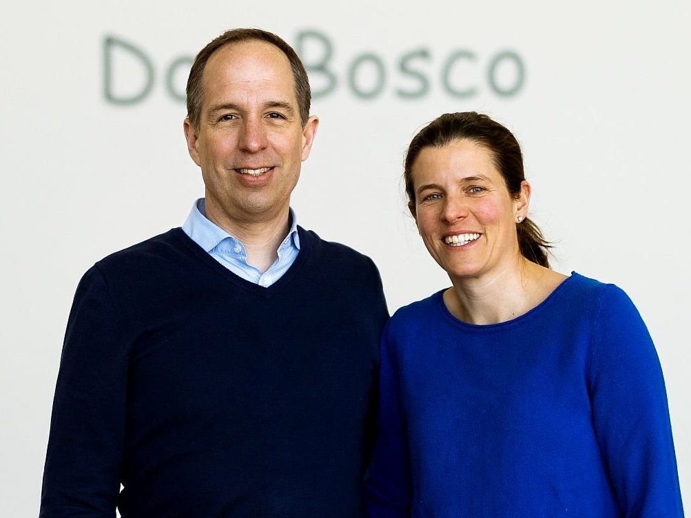 Ein lächelnder Mann im blauen Pullover und eine Frau im blauen Oberteil stehen nebeneinander vor einer weißen Wand mit 'Do Bosco'-Schriftzug.