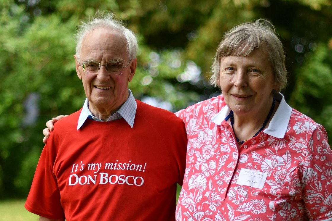 Lächelndes älteres Paar im Freien. Mann trägt ein 'It's my mission! DON BOSCO' T-Shirt, Frau ein florales Poloshirt mit Namensschild.
