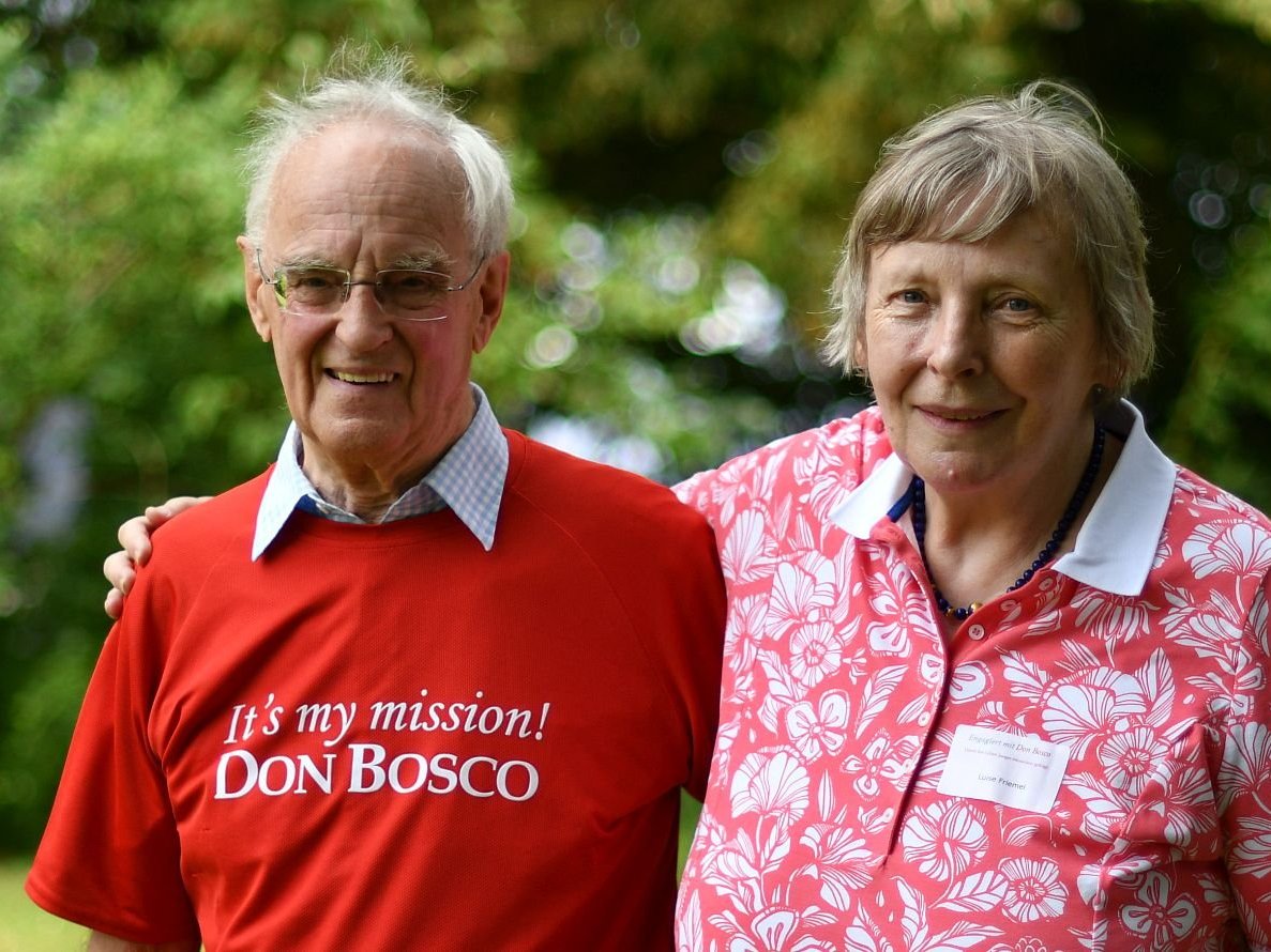 Lächelndes älteres Paar im Freien. Mann trägt ein 'It's my mission! DON BOSCO' T-Shirt, Frau ein florales Poloshirt mit Namensschild.
