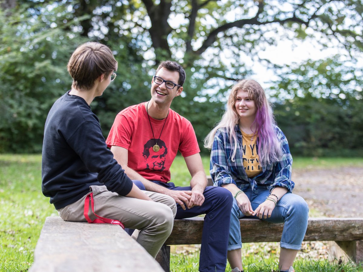 Drei vielfältige junge Leute lächeln und unterhalten sich auf einer Holzbank im Park, umgeben von Grün.