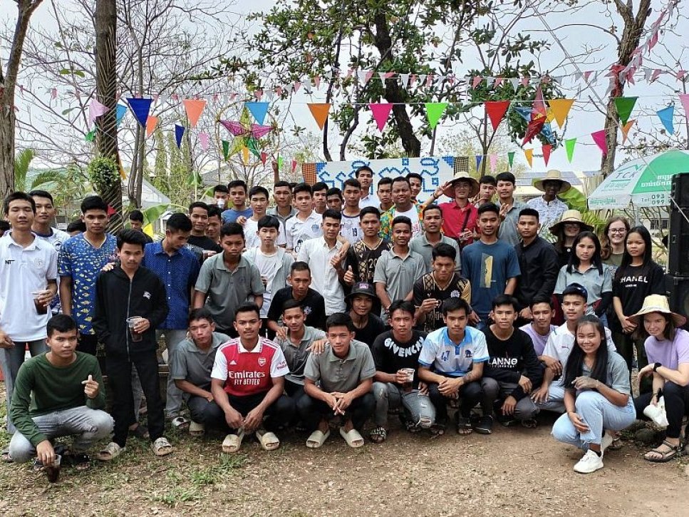 Eine große Gruppe junger Menschen, hauptsächlich Männer, posiert draußen unter bunten Wimpeln für ein Gruppenfoto.