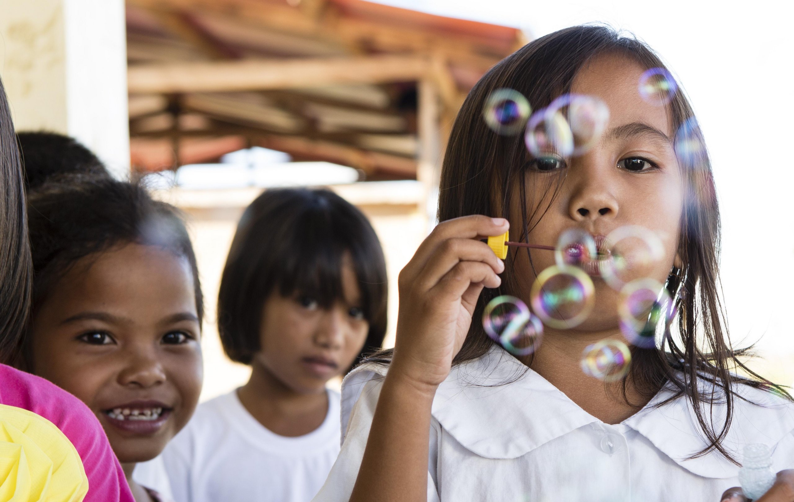 Mädchen in weißer Schuluniform bläst lächelnd bunte Seifenblasen, andere Kinder sind im Hintergrund sichtbar.