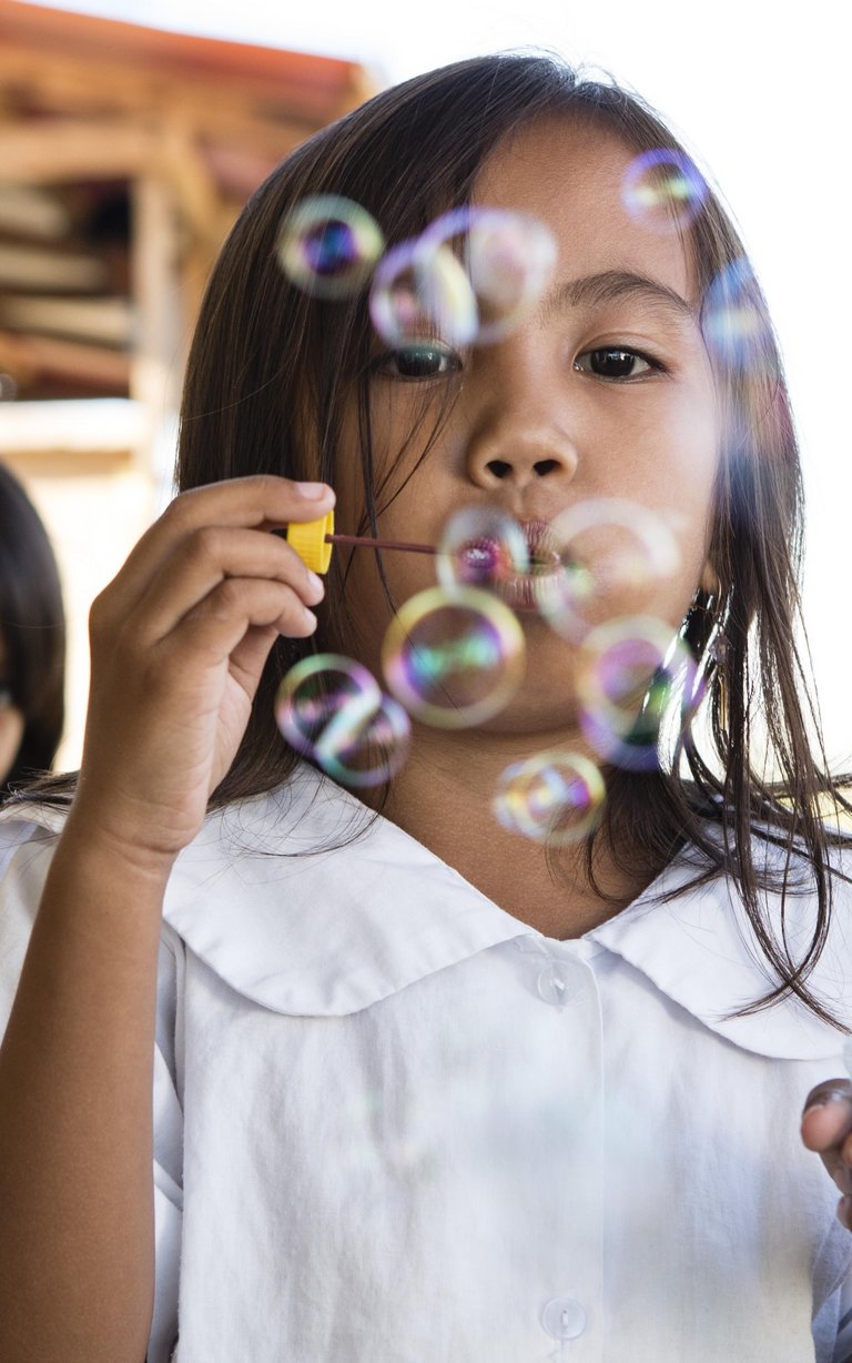 Mädchen in weißer Schuluniform bläst lächelnd bunte Seifenblasen, andere Kinder sind im Hintergrund sichtbar.