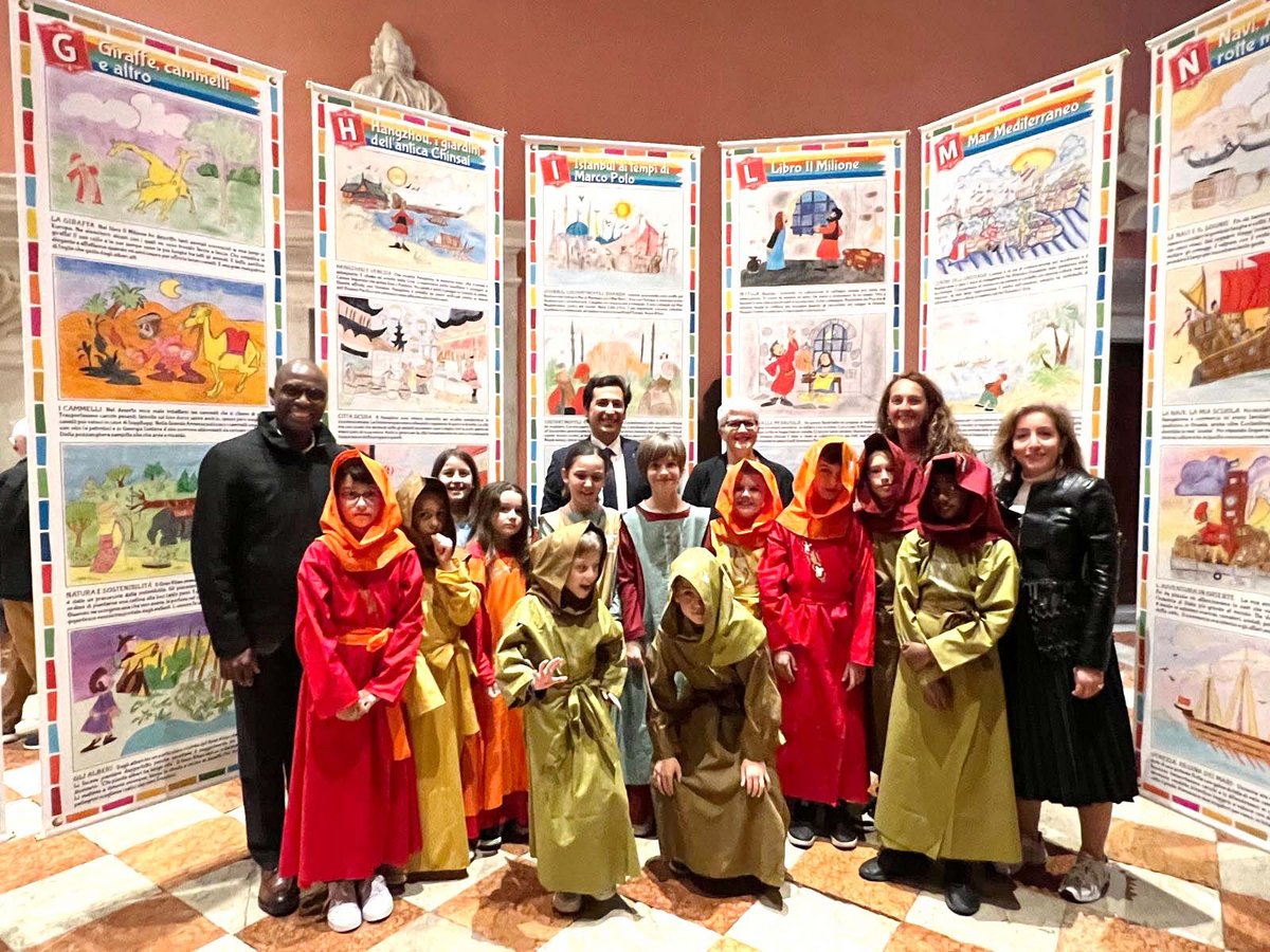 Kinder in roten, orangen und olivgrünen historischen Gewändern posieren mit Erwachsenen vor bunten Plakaten über Marco Polo.