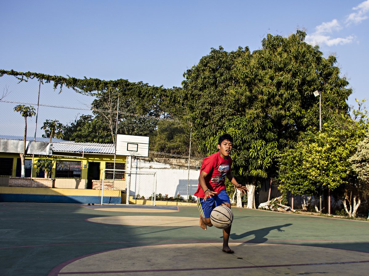 Barfüßiger junger Mann in rotem Shirt dribbelt Basketball auf Außenplatz. Korb, gelbes Gebäude, grüne Bäume im Hintergrund.