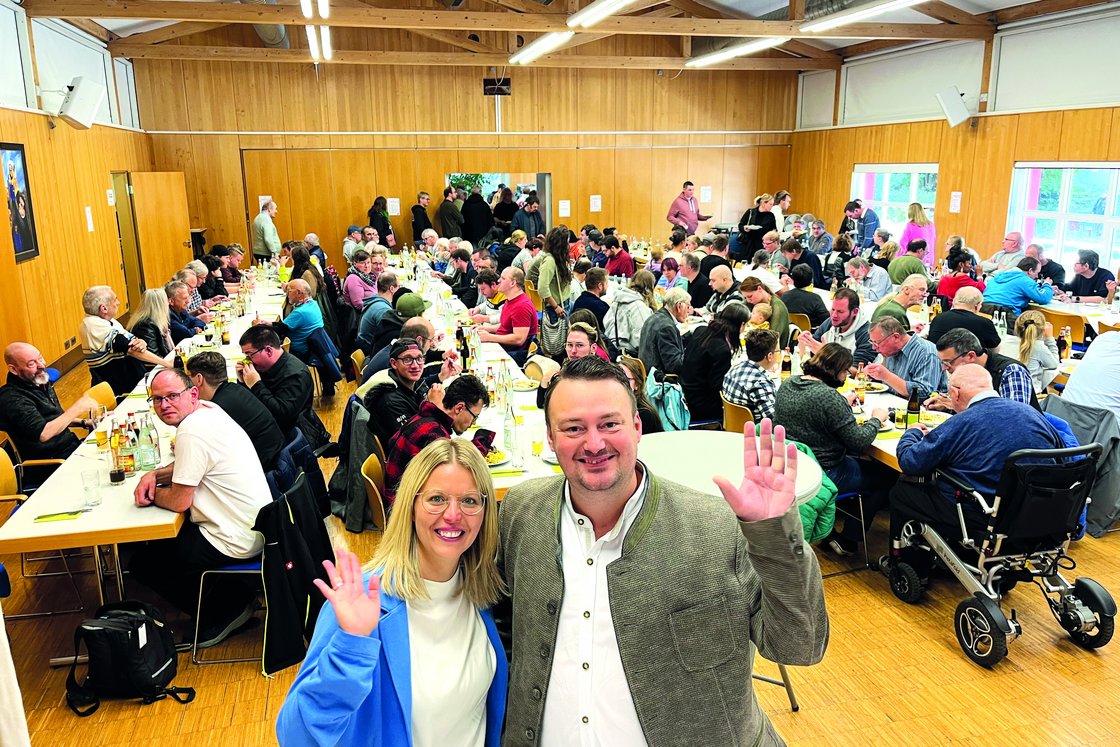Eine große Gruppe von Menschen genießt eine Mahlzeit bei einer Indoor-Veranstaltung, mit einem lächelnden Paar, das in die Kamera winkt.