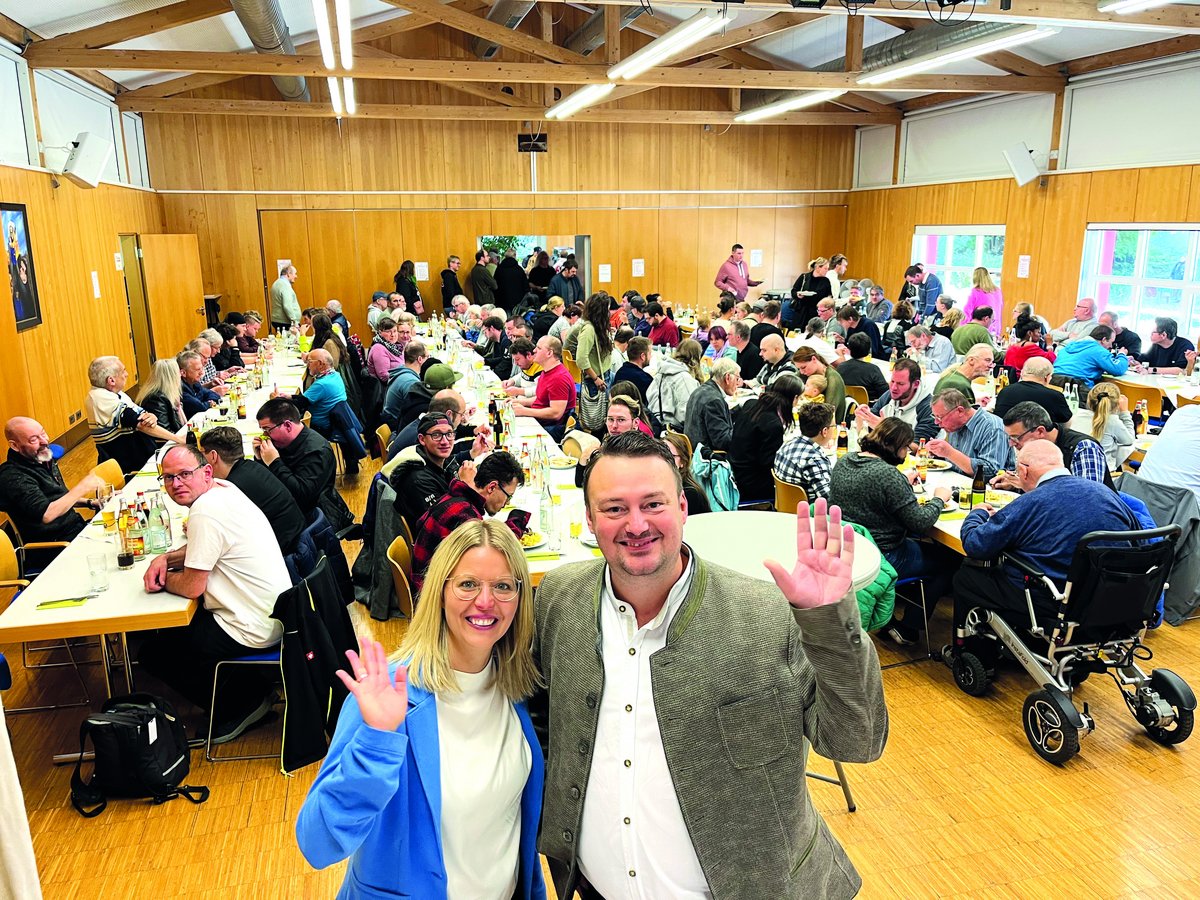 Eine große Gruppe von Menschen genießt eine Mahlzeit bei einer Indoor-Veranstaltung, mit einem lächelnden Paar, das in die Kamera winkt.