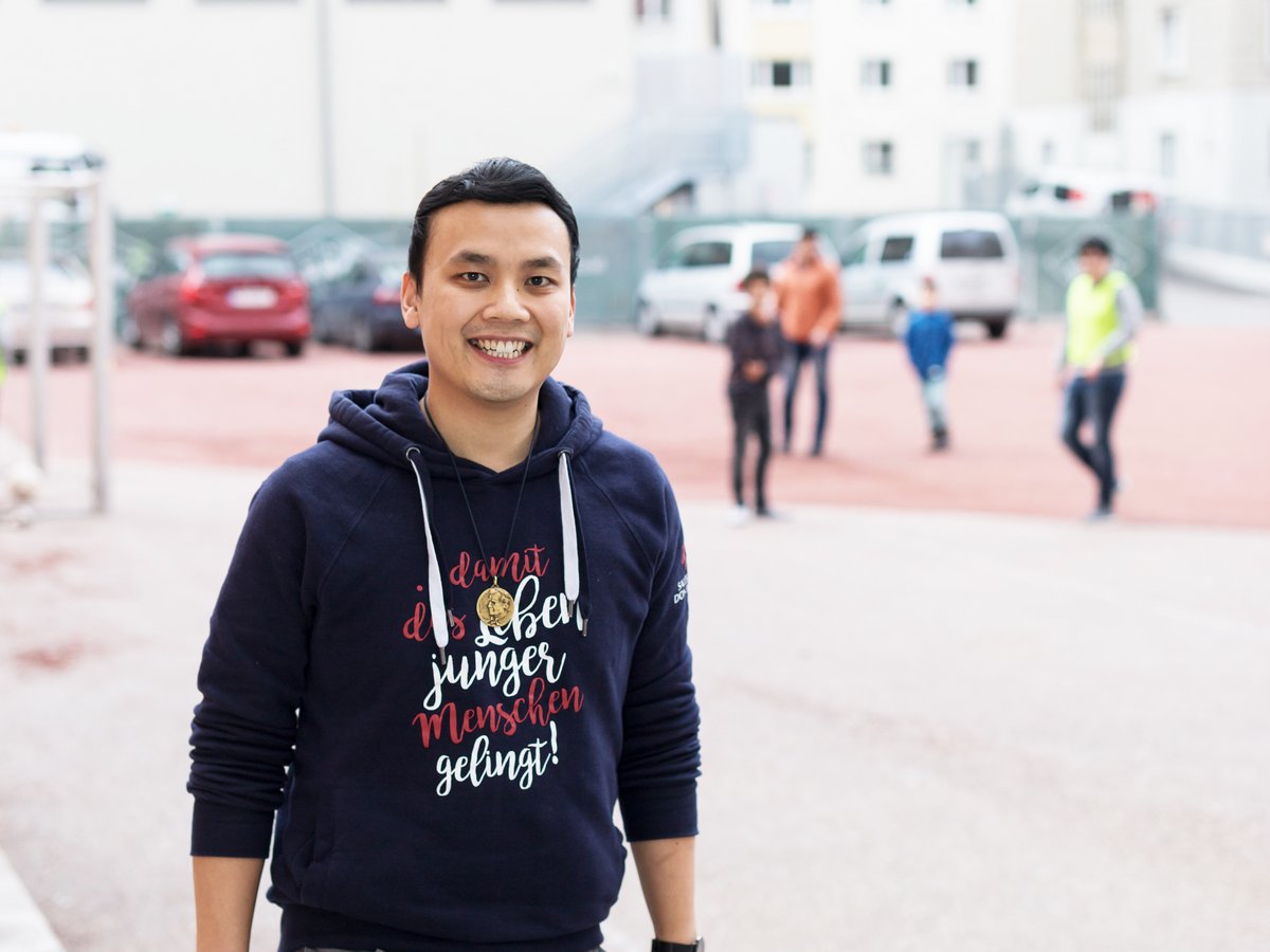 Lächelnder asiatischer Mann in blauem Hoodie mit deutschem Text 'damit das Leben junger Menschen gelingt!', im Freien.