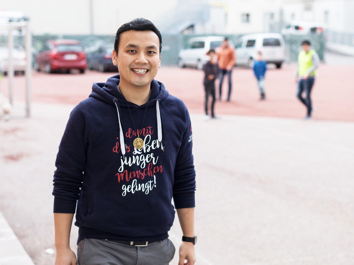 Lächelnder asiatischer Mann in blauem Hoodie mit deutschem Text 'damit das Leben junger Menschen gelingt!', im Freien.