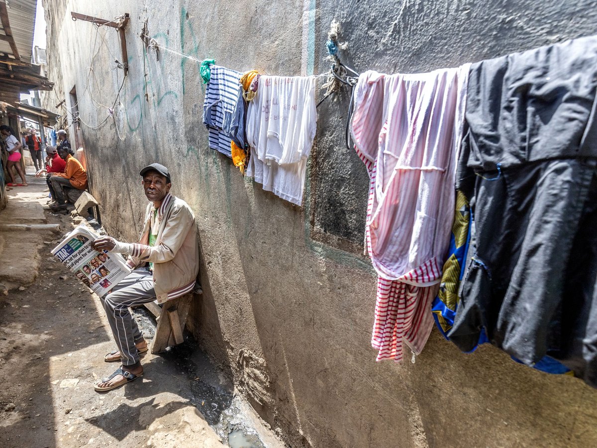 Mann liest Zeitung in einer engen, sonnigen Slum-Gasse, mit Wäsche auf der Leine und anderen Menschen im Hintergrund.