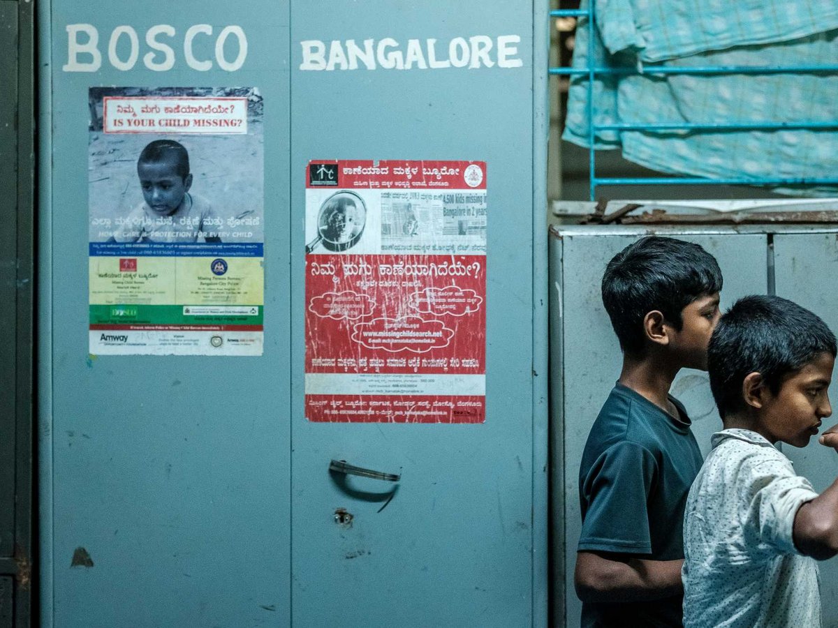 Zwei Jungen stehen neben einem Metallschrank mit Plakaten über vermisste Kinder. Einer trinkt aus einem Metallbecher in einer urbanen Szene.