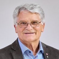 Schützner-Norbert-SDB Kopfaufnahme eines lächelnden Mannes mittleren Alters mit grauen Haaren und Brille, ein Hemd tragend, vor einem unscharfen grünen Hintergrund.