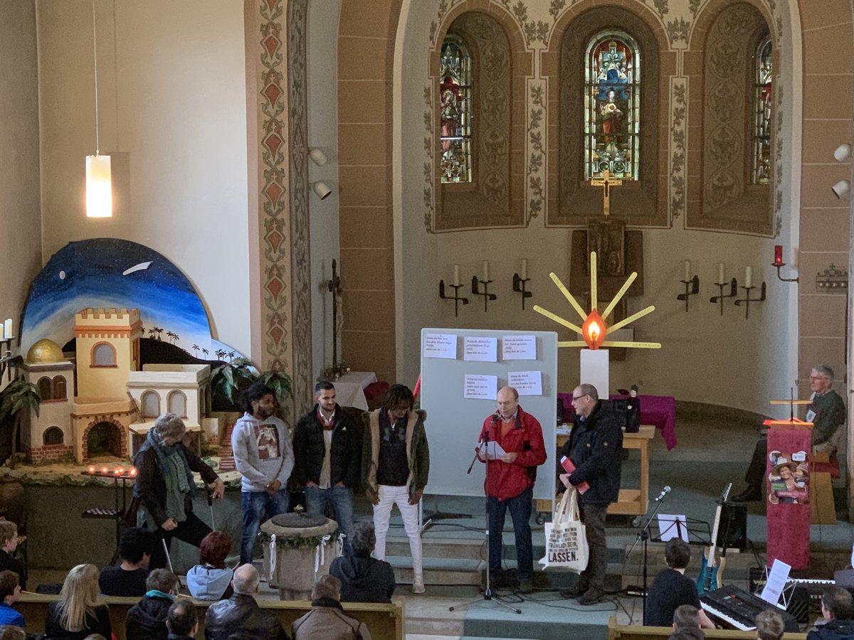 Menschen in einer Kirche, junge Männer und Redner vor Taufbecken und festlicher Dekoration, Publikum in Bänken.