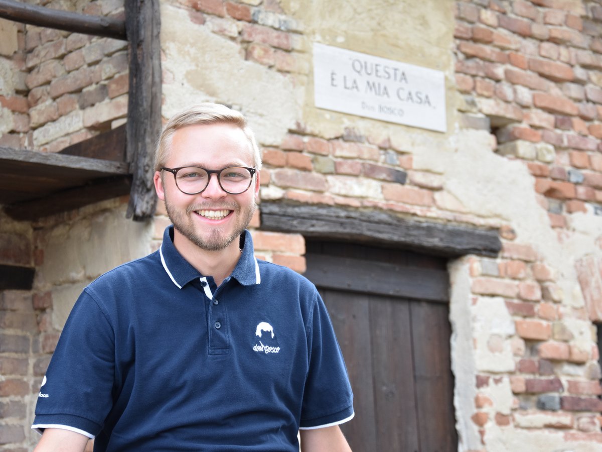 Ein lächelnder Mann in einem Don Bosco Poloshirt steht vor einem alten Backsteingebäude mit einer "Questa è la mia casa" Tafel.