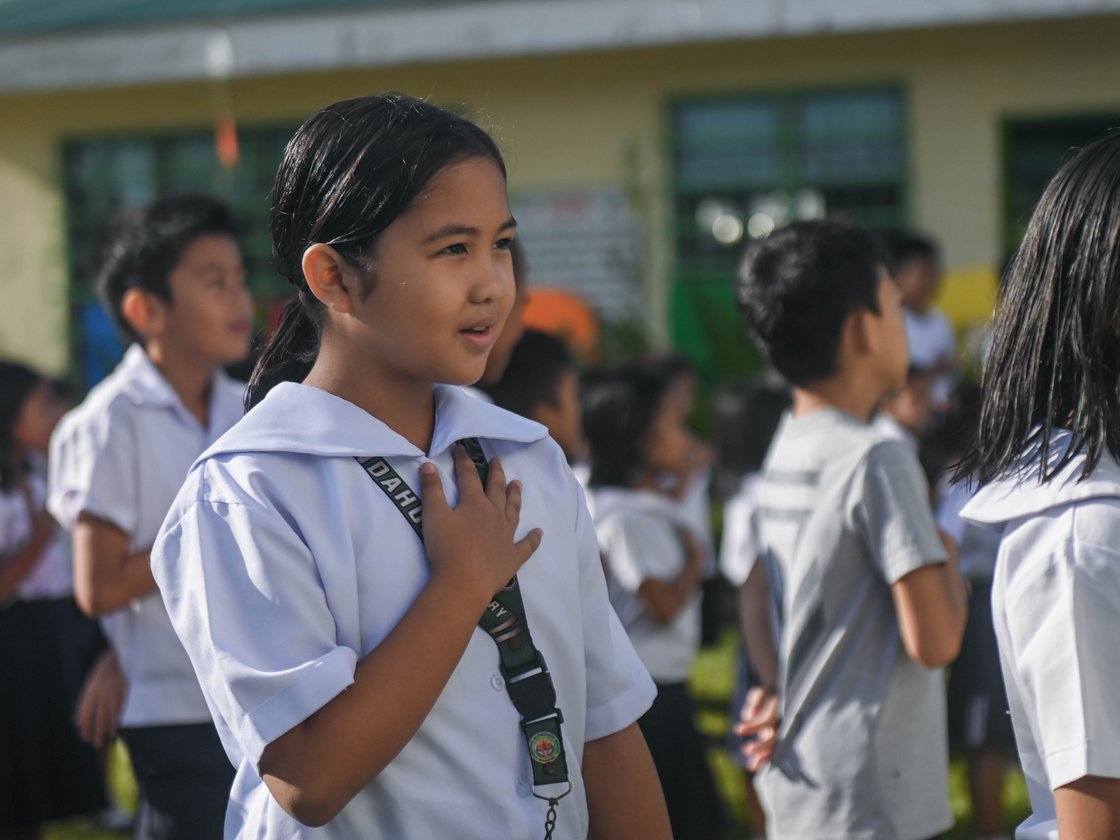 Eine Schülerin in weißer Uniform mit der Hand auf ihrer Brust, die draußen mit anderen Schülern steht.