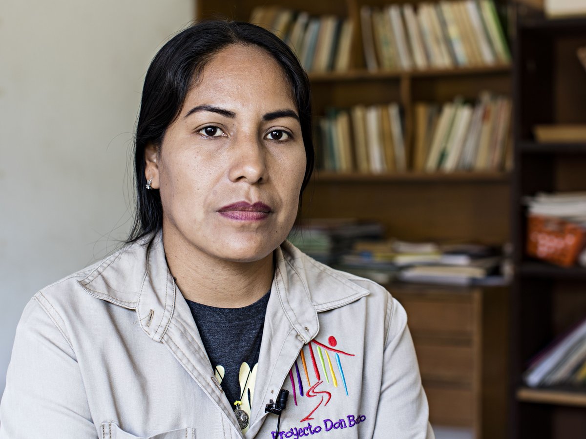 Porträt einer Frau mit dunklen Haaren und ernstem Blick, die eine Jacke mit "Proyecto Don Bobo"-Logo trägt, vor Bücherregalen.