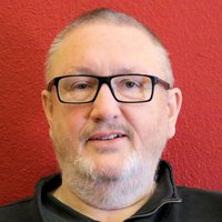 Ein lächelnder Mann mit Brille und weißem Bart trägt eine schwarze Jacke vor rotem Hintergrund.