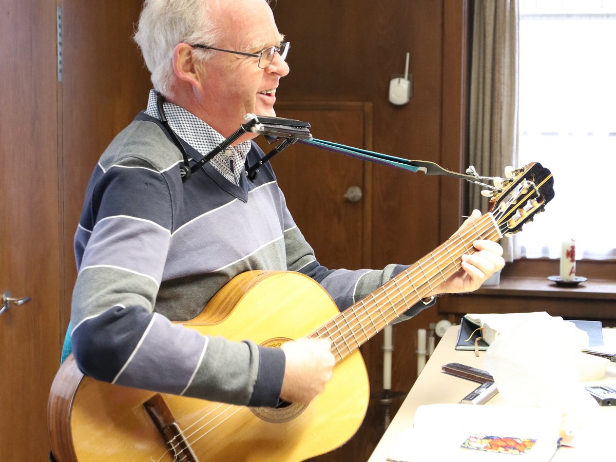 Ein lächelnder Mann mit Brille und gestreiftem Pullover spielt gleichzeitig Akustikgitarre und Mundharmonika in einem Raum.