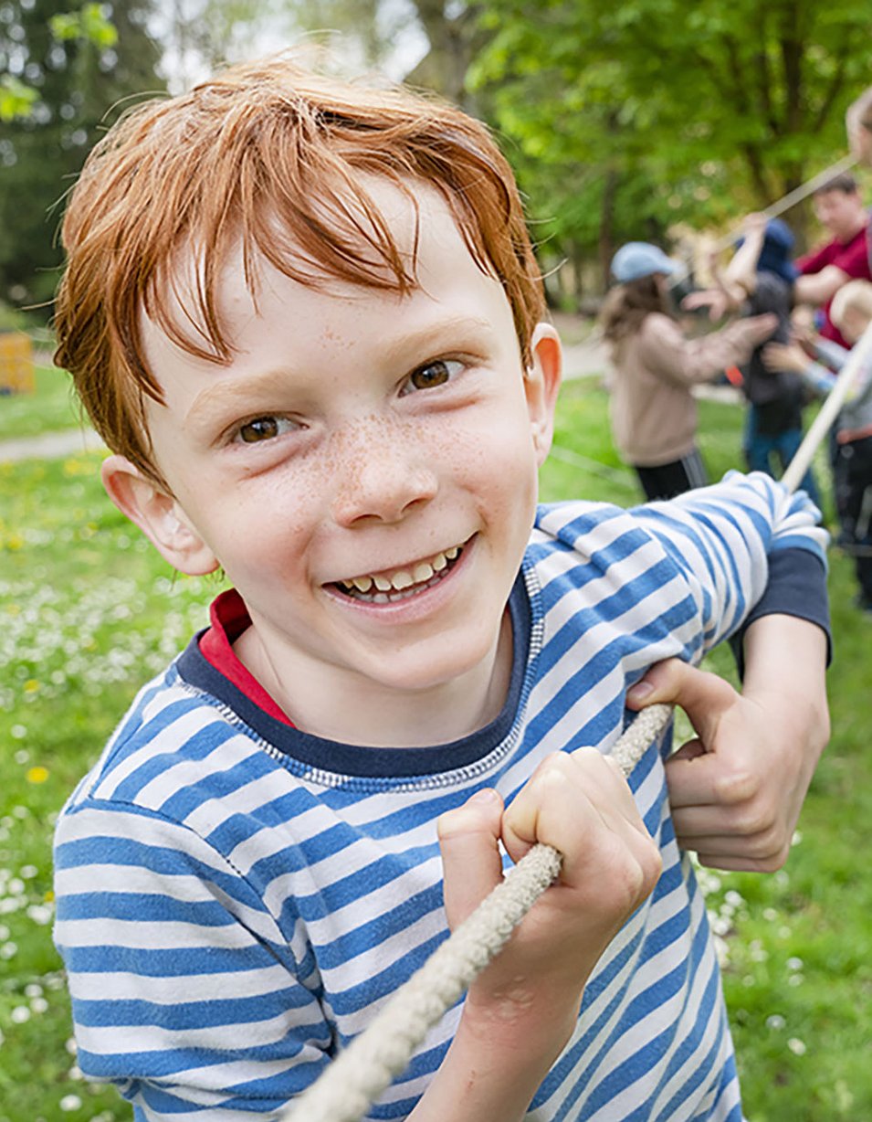 Fröhlicher rothaariger Junge hält ein Seil; im Hintergrund andere Kinder bei einem Seilparcours im Freien.