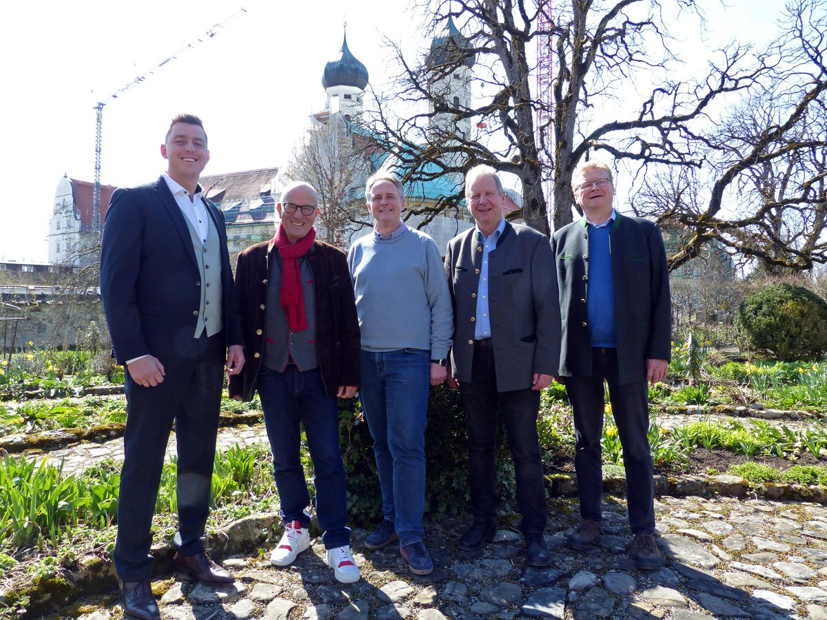 Fünf lächelnde Männer in Tracht und Anzügen in einem Garten mit historischer Kirche und Kran im Hintergrund.