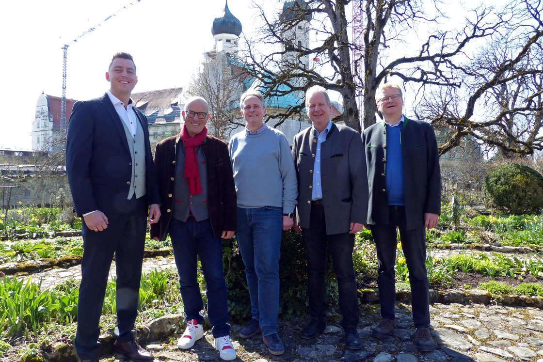 Fünf lächelnde Männer in Tracht und Anzügen in einem Garten mit historischer Kirche und Kran im Hintergrund.