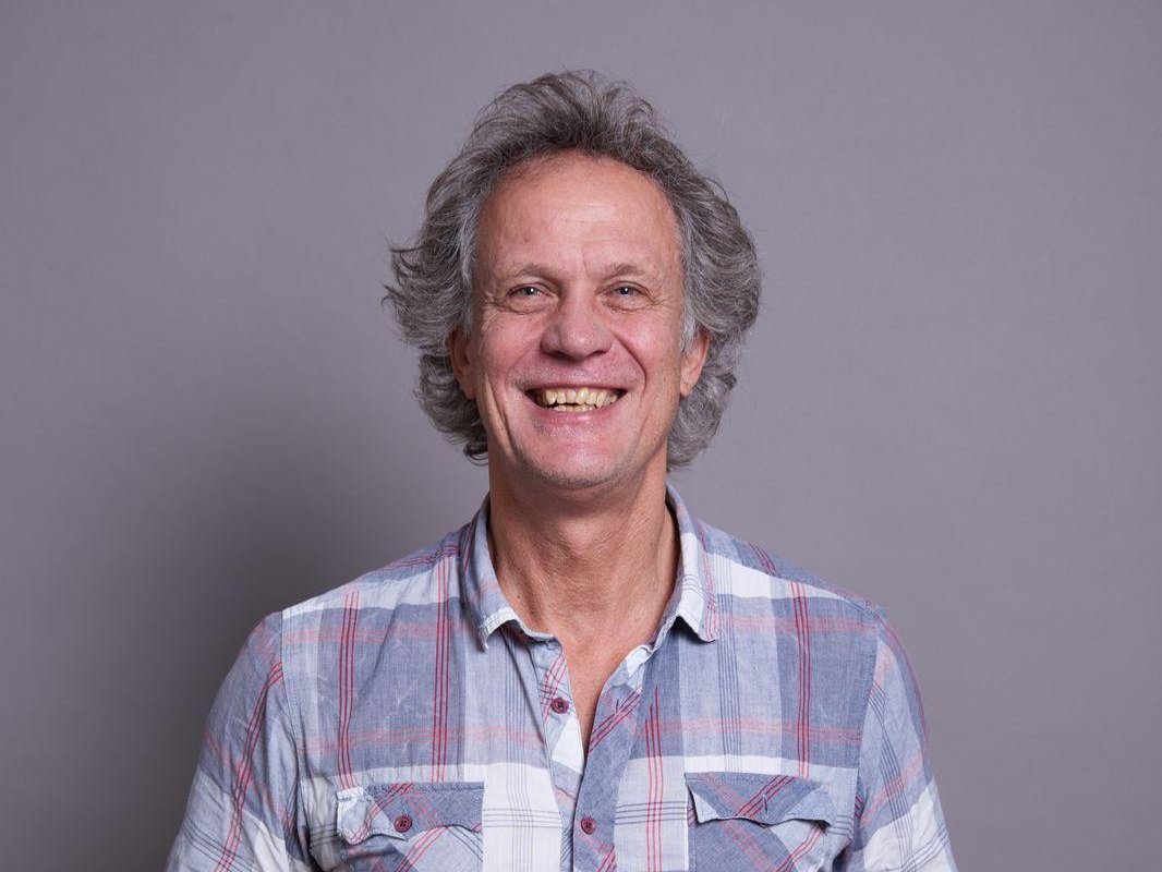 Ein lächelnder Mann in einem karierten Hemd hält eine Modellbahnweiche und zeigt darauf, vor grauem Hintergrund.