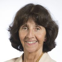 Lächelnde ältere Frau mit dunkelbraunem Haar und hellen Augen, trägt ein helles Oberteil und eine Perlenkette, vor weißem Hintergrund.