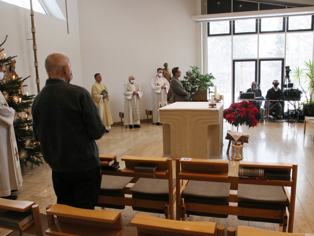Kirchlicher Gottesdienst mit Priestern (Masken), Weihnachtsbaum und Medientechnik vor verschneitem Fenster.