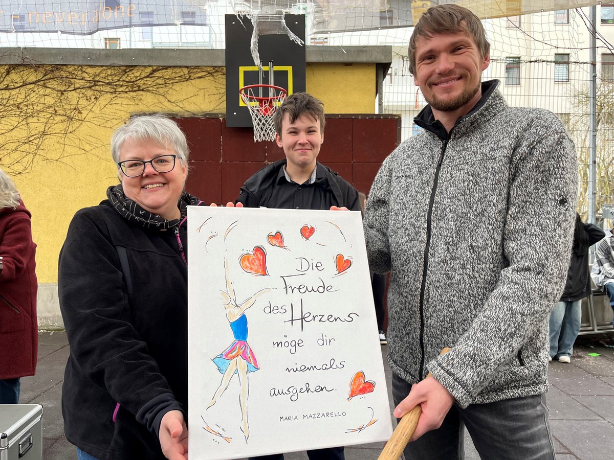 Drei lächelnde Personen halten ein Gemälde mit Herzen, einer Tänzerin und dem Text "Die Freude des Herzens möge dir niemals ausgehen."