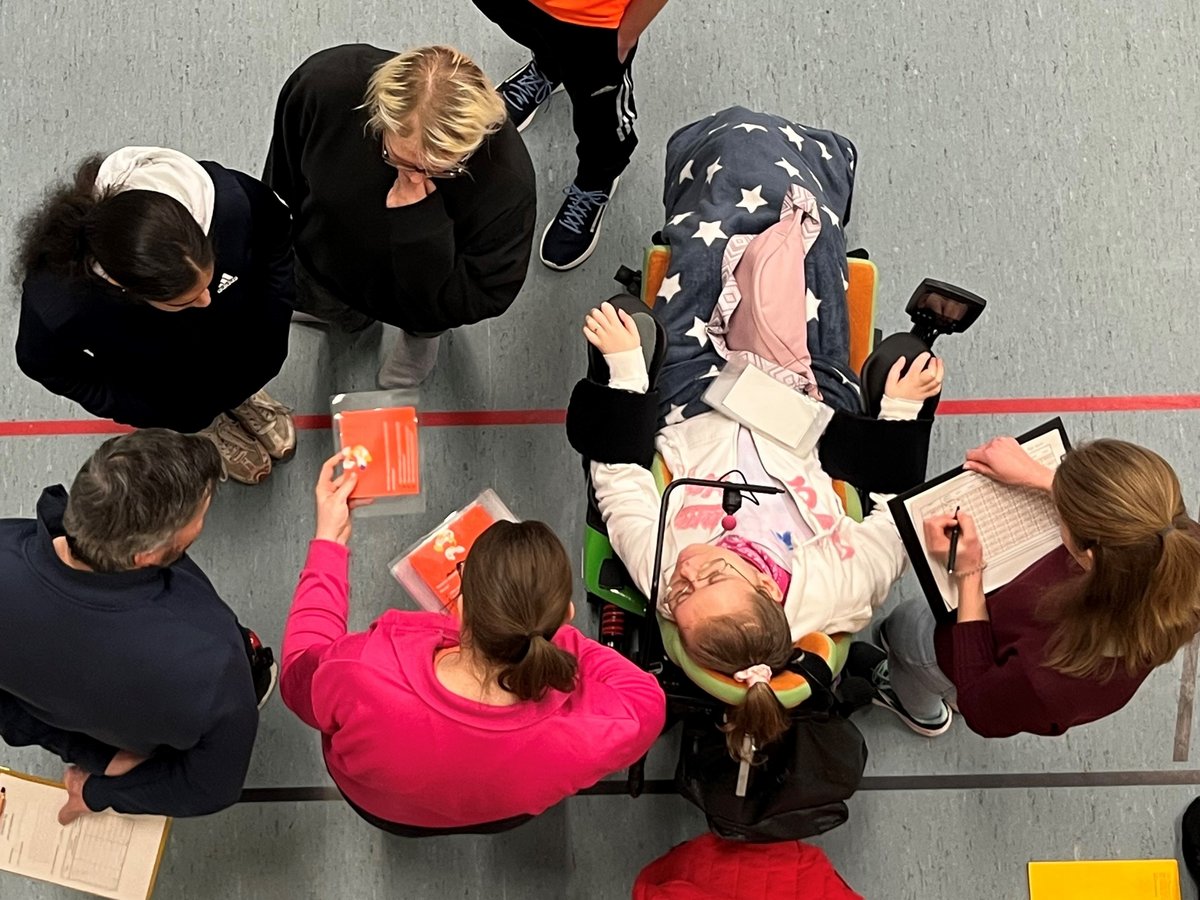 Draufsicht auf eine Person im Rollstuhl, die an einer Aktivität teilnimmt, umgeben von Leuten mit Papieren und Klemmbrettern.