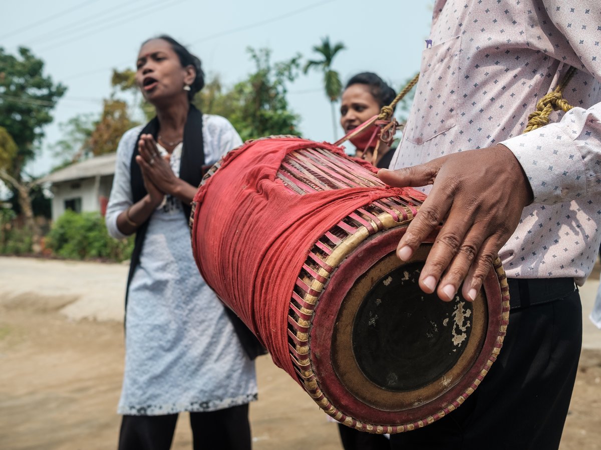 Eine Person hält eine rot umwickelte Trommel, ihre Hand ruht auf der Oberfläche. Im unscharfen Hintergrund singt eine Frau mit gefalteten Händen.