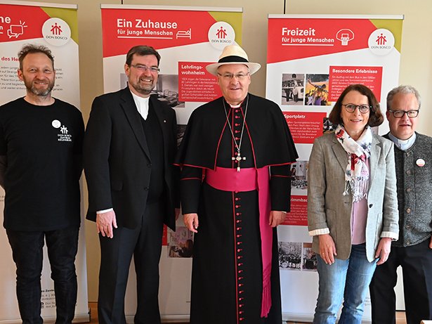 Gruppe von sechs Personen, darunter ein Bischof mit Hut und weitere Geistliche, vor Don Bosco Bannern.