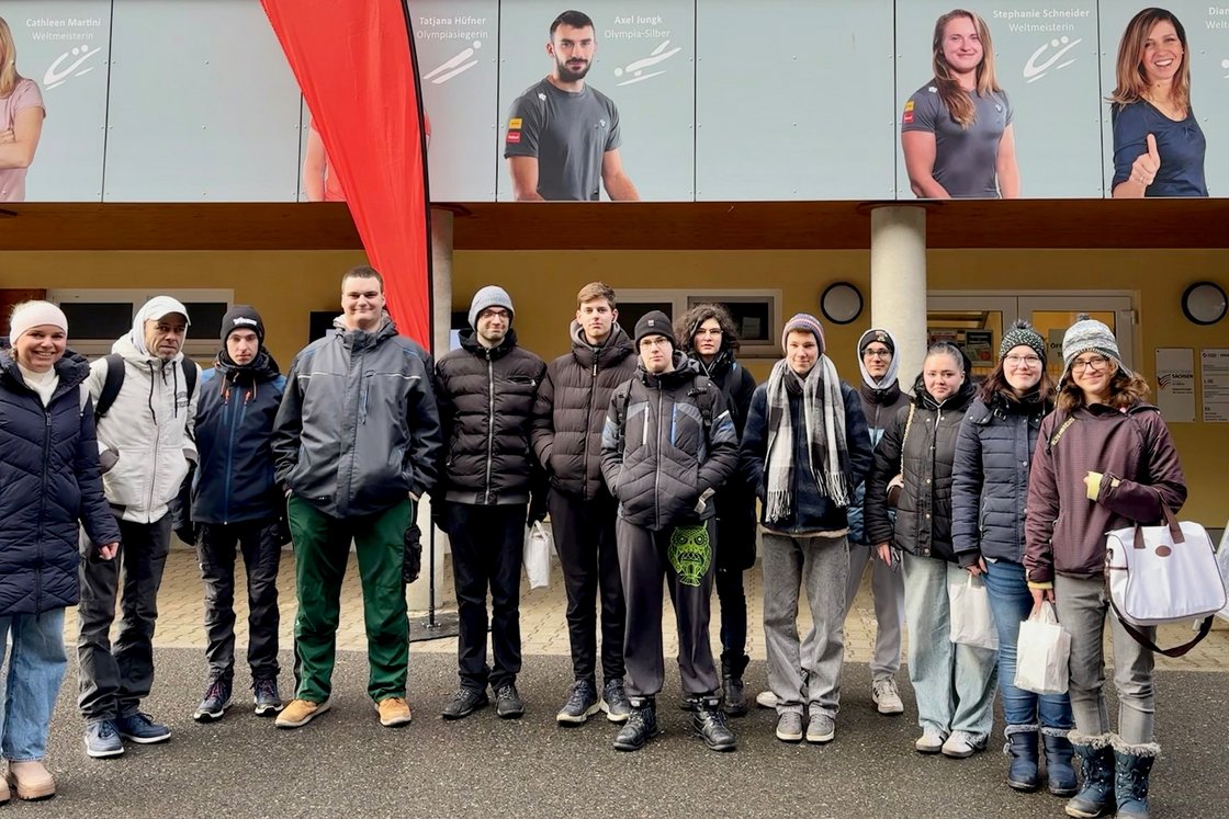 Eine Gruppe junger Menschen und Erwachsener in Winterkleidung steht lächelnd vor einem Gebäude mit Postern von Sportlern.