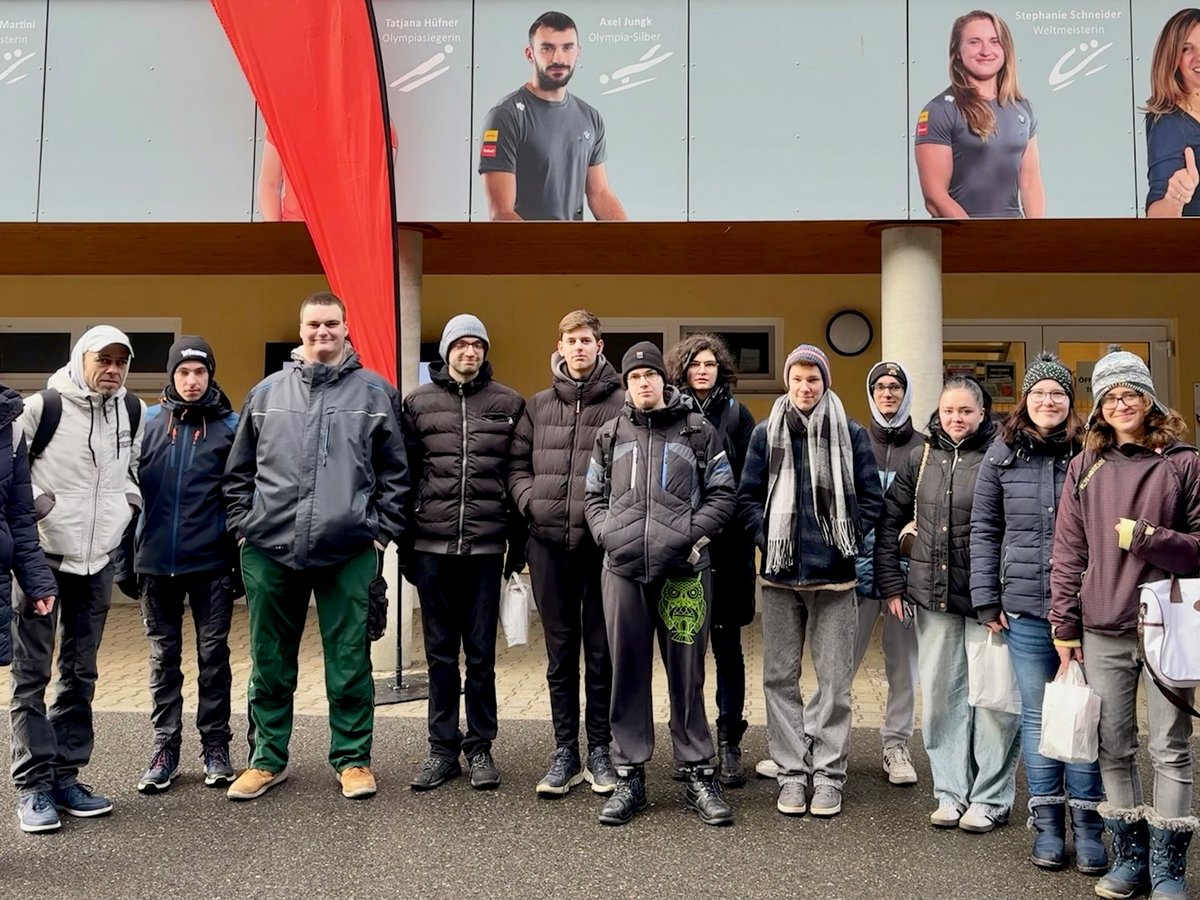 Eine Gruppe junger Menschen und Erwachsener in Winterkleidung steht lächelnd vor einem Gebäude mit Postern von Sportlern.